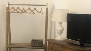 Una habitación con un aparador de madera, un televisor encima, una lámpara al lado, un estante de madera con ropa y una cesta de mimbre.