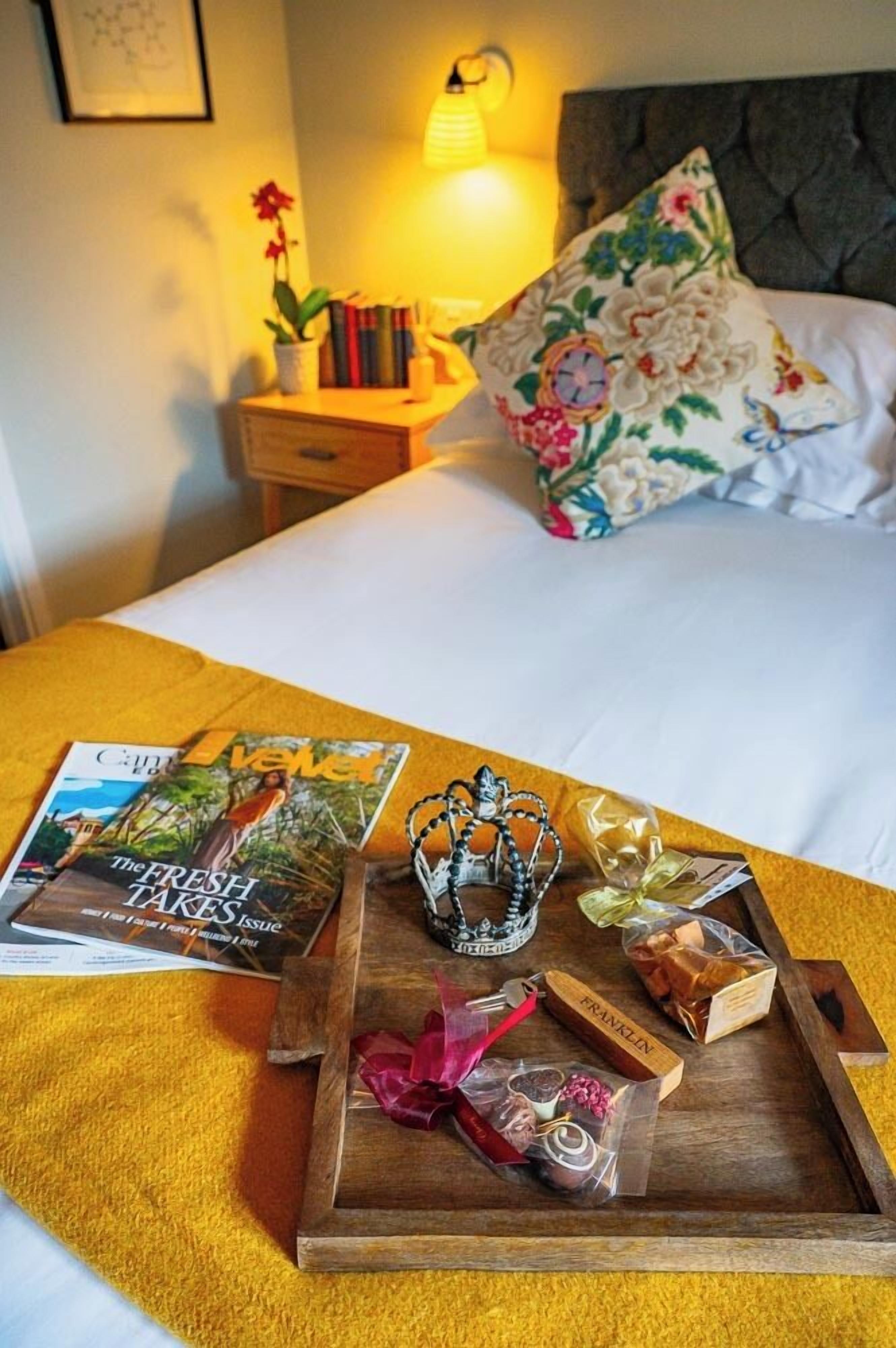 Una cama bien tendida con un cojín floral, una mesita de noche con libros y una lámpara, y una bandeja con bombones y una corona.