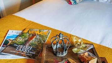 Una cama bien tendida con un cojín floral, una mesita de noche con libros y una lámpara, y una bandeja con bombones y una corona.