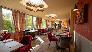 Een restaurant met meerdere eettafels en stoelen, grote ramen met gordijnen, en diverse hangende hanglampen.