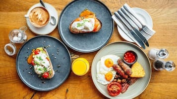 Ein Frühstückstisch mit verschiedenen Speisen, darunter Toast mit Sahne, Eier und Würstchen, dazu eine Tasse Kaffee und ein Glas Orangensaft.