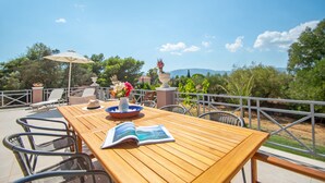 Una mesa de madera con un libro, un florero con flores y una estatua decorativa en un patio al aire libre con sillas y una barandilla.