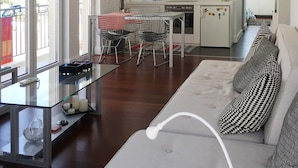 Uma sala de estar moderna com uma mesa de centro de vidro, um sofá cinza com almofadas estampadas e uma área de cozinha com armários brancos e uma mesa de jantar com cadeiras.