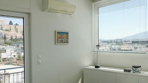 Um quarto com cama, radiador, janela com persianas, quadro na parede e varanda com vista para a paisagem urbana.