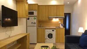 A compact kitchen with wooden cabinets, a microwave, a refrigerator, and a washing machine. There is a sofa and a TV mounted on the wall.