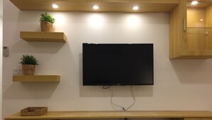 A room with a mounted flat-screen TV, wooden shelves with plants, a wooden desk with drawers, and a remote control on the desk.
