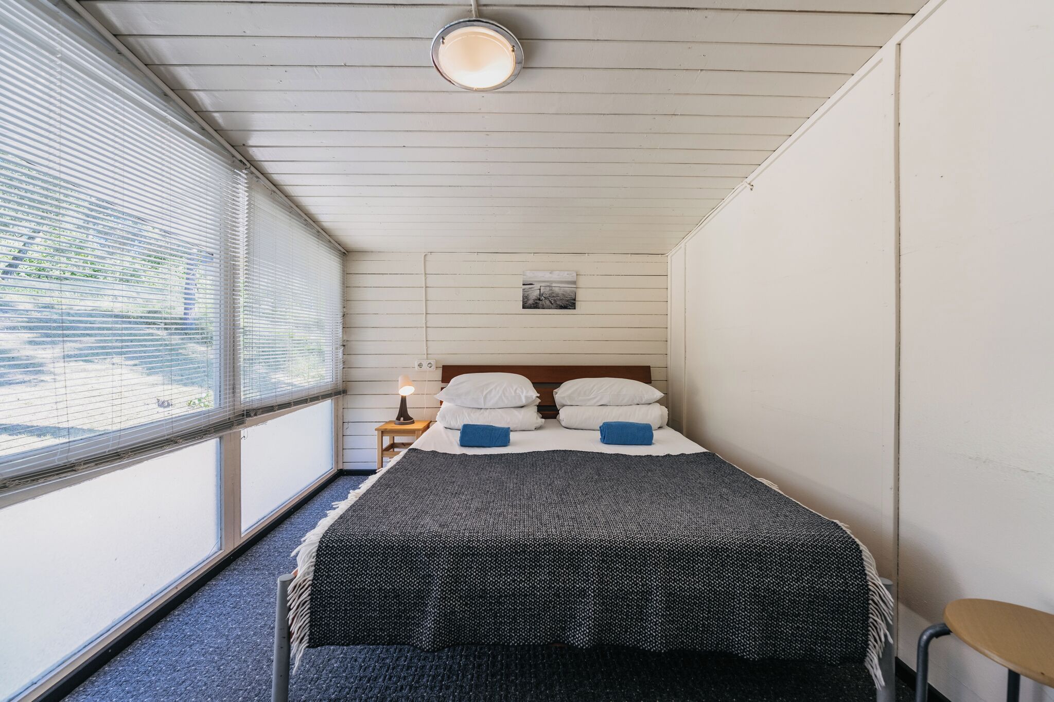 A single bed with a grey and black patterned bedspread, white pillows, and two blue towels. There is a small bedside table with a lamp, a framed picture on the wall, and a window with blinds.