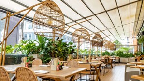 A restaurant with wooden tables and chairs, large windows, and hanging wicker lamps.