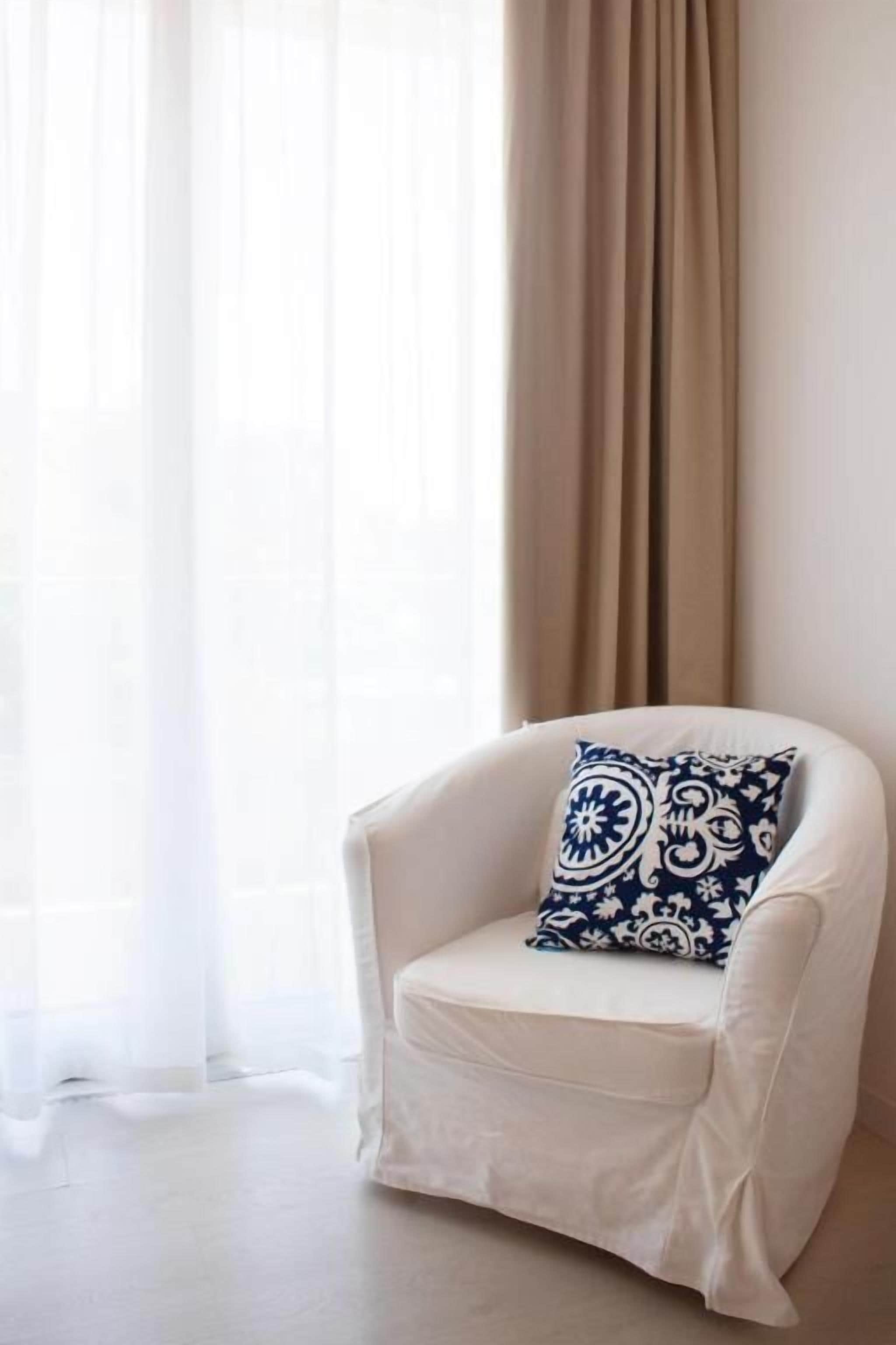 A white armchair with a patterned cushion in a room with a sheer curtain and a solid-colored drape.