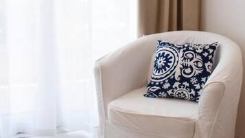 A white armchair with a patterned cushion in a room with a sheer curtain and a solid-colored drape.