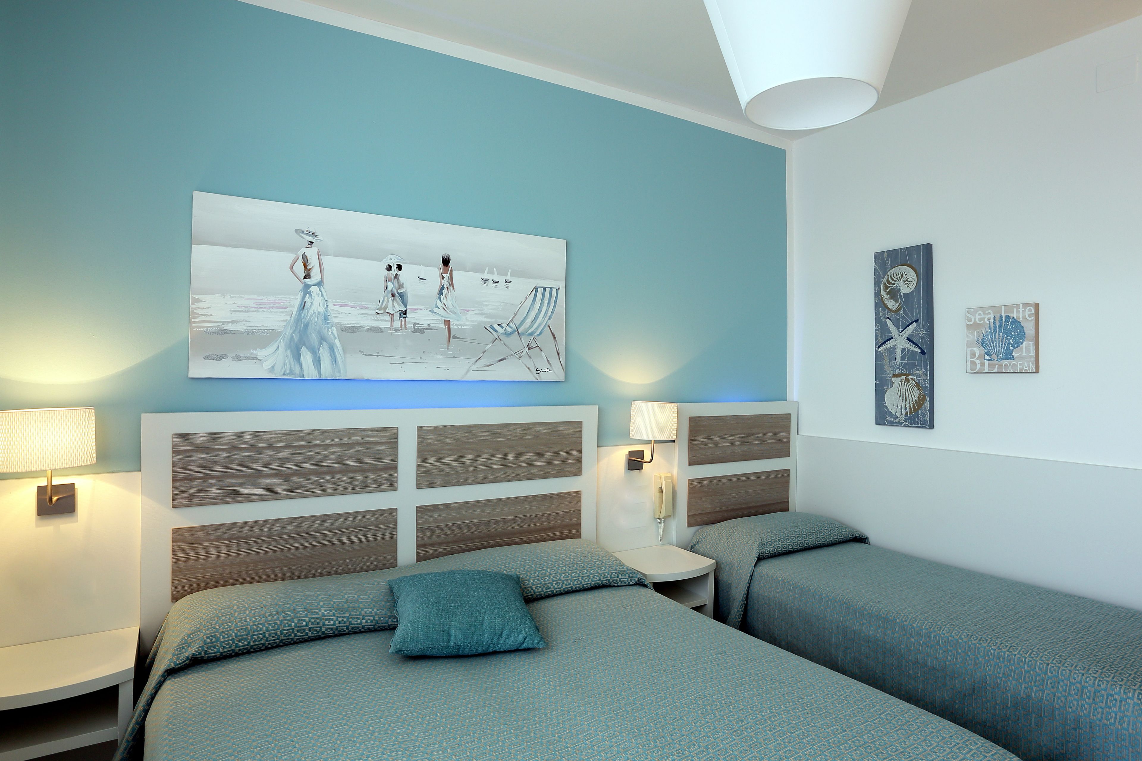 Ein Schlafzimmer mit zwei Betten, einem Nachttisch mit Lampe, einem Wandtelefon und einem Gemälde mit Strandmotiv.