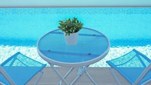Ett runt glasbord med en krukväxt i mitten, två blå fåtöljer och en pool med blå kakelplattor i bakgrunden.