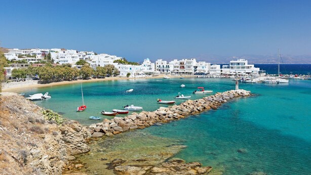 A coastal town with white buildings, a rocky breakwater, and boats in the water.
