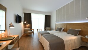 Een hotelkamer met twee bedden, een bureau met lamp, een stoel, een aan de muur bevestigde televisie en een groot raam met gordijnen.