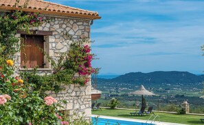 Un bâtiment en pierre doté d’une piscine, entouré d’une végétation luxuriante et offrant une vue sur des collines lointaines.