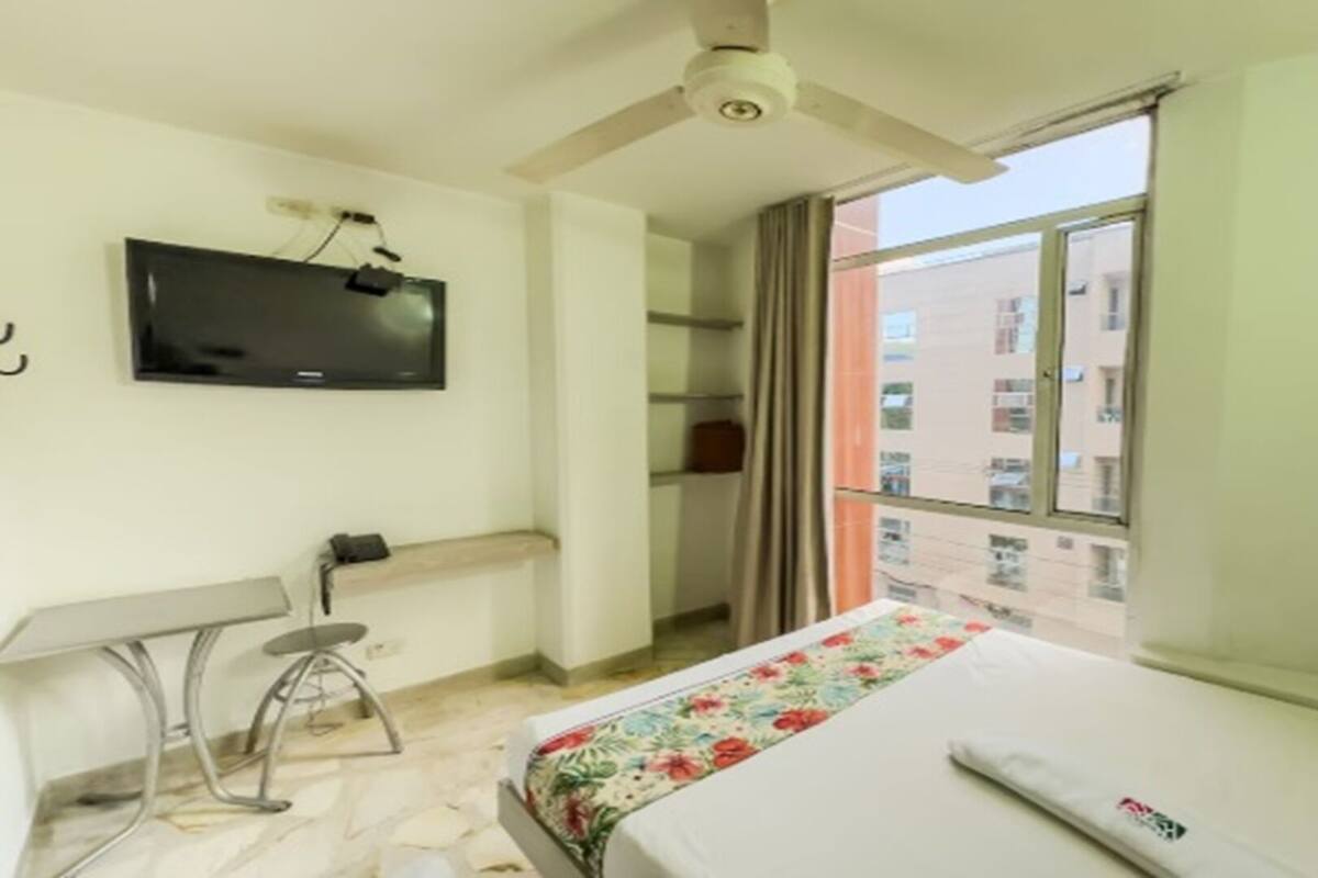 Habitació d'hotel amb un llit, una taula de treball, una cadira, un televisor i una finestra amb vista a edificis.