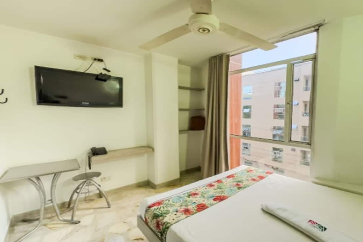 A hotel room with a bed, a desk, a chair, a television, and a window with a view of buildings.