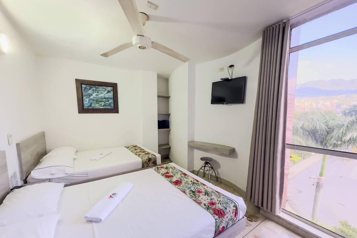 Habitació d'hotel amb dos llits, un ventilador de sostre, televisió, finestra amb vistes i un petit tamboret.