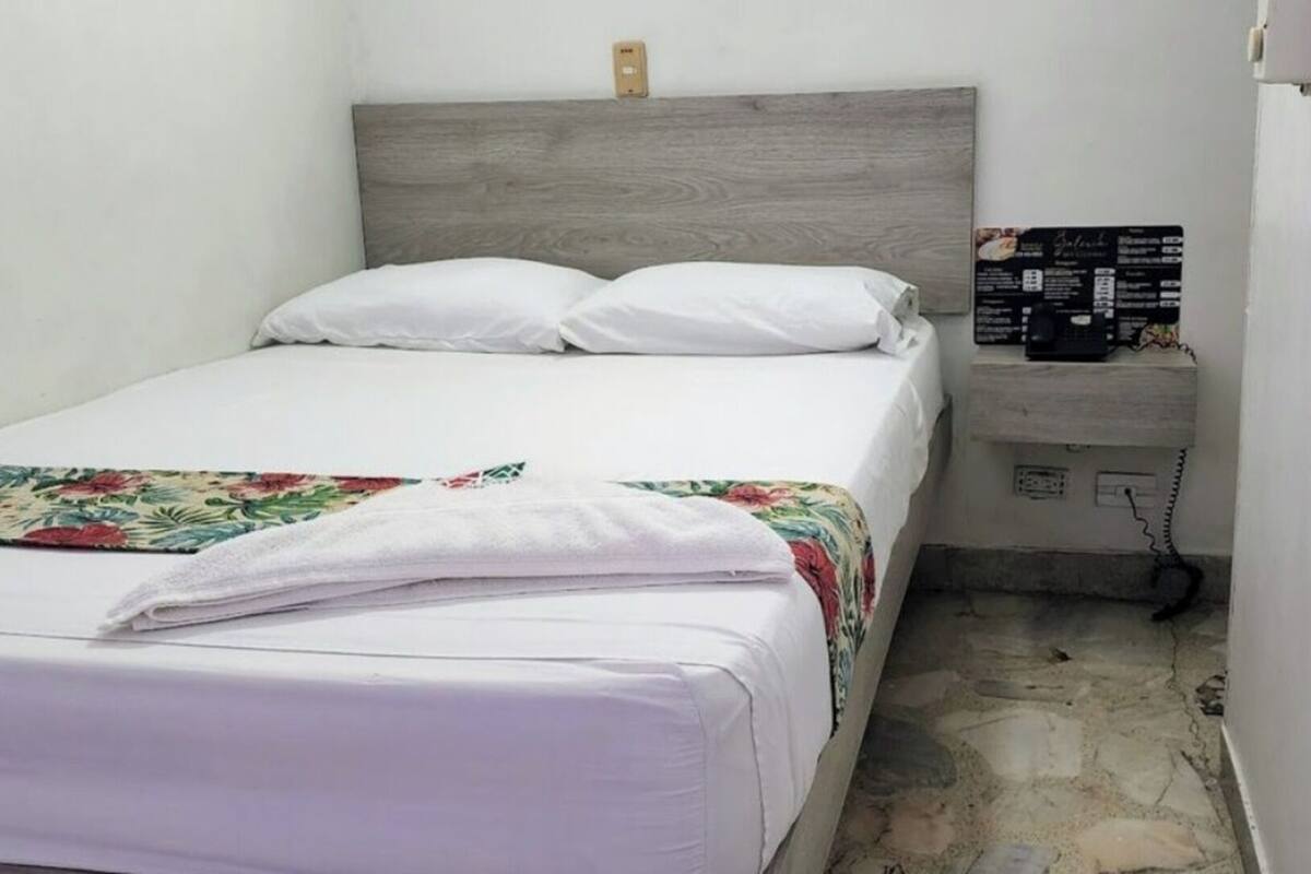 Un llit petit i ben fet amb roba de llit blanca i una manta amb estampat floral, un capçal de fusta, una taula de nit amb un objecte negre i un sòl amb motius de pedra.