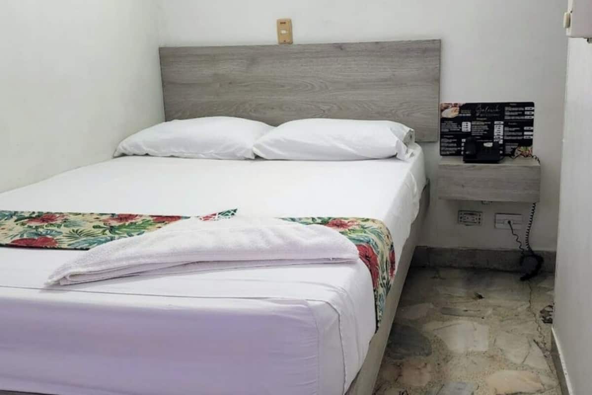 A small, neatly made bed with white linens and a floral-patterned blanket, a wooden headboard, a bedside table with a black object, and a stone-patterned floor.
