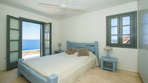 Een slaapkamer met een bed, nachtkastje, lamp, plafondventilator en uitzicht op zee door open deuren.