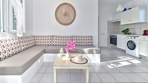 A modern kitchen with a built-in washing machine, a dining area with a table and chairs, and a patterned bench. - Poseidon | 2 Bedroom Family Apartment with Sea View and Patio, Cleopatra Homes Paros (Paros)