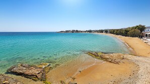 A coastal beach with clear turquoise waters, sandy shore, and buildings in the background. - Poseidon | 2 Bedroom Family Apartment with Sea View and Patio, Cleopatra Homes Paros (Paros)