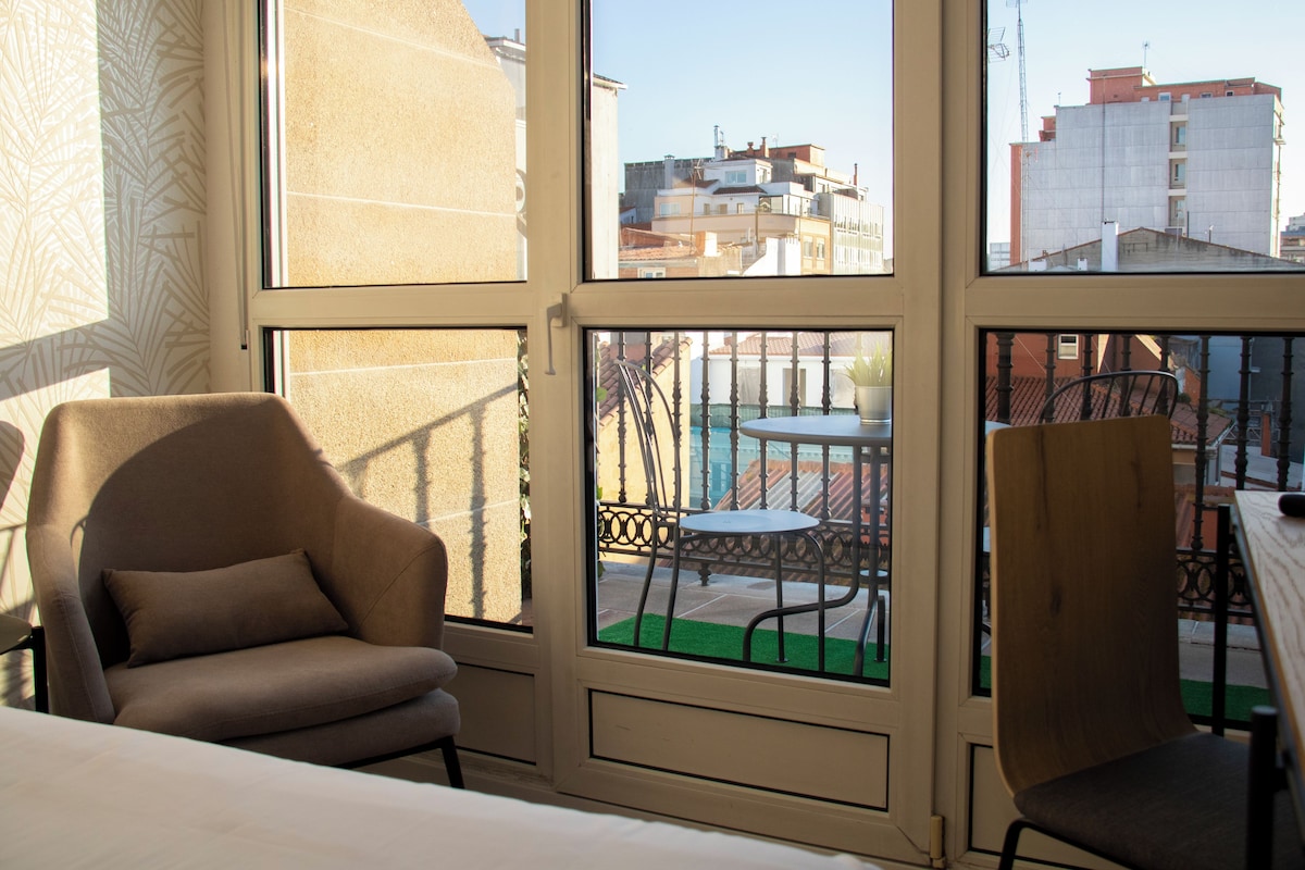 A room with a large window, a chair, a small table, and a view of buildings outside.