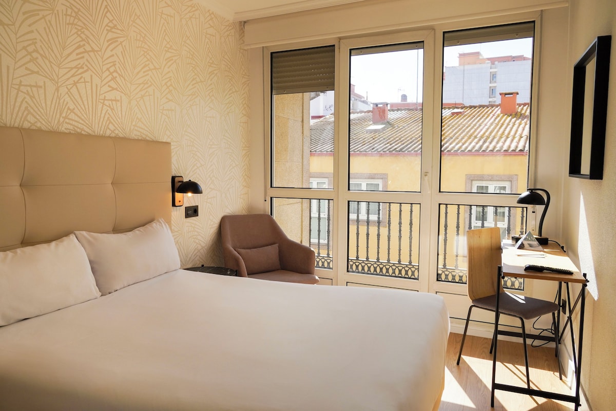 A hotel room with a large bed, a desk with a chair, and a balcony with a view of buildings.