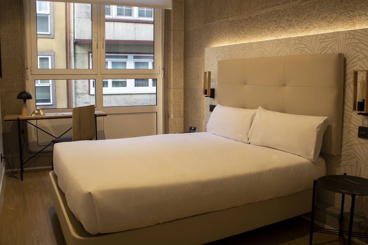 Una cama bien tendida en una habitación de hotel con un ventanal grande, un escritorio con una lámpara y una mesita.