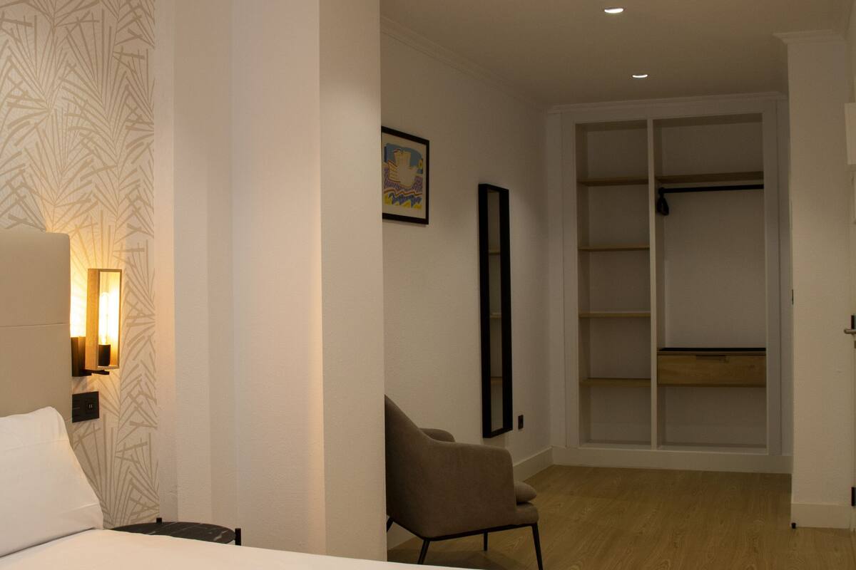 Habitación de hotel con cama, una silla, armario e iluminación en la pared.