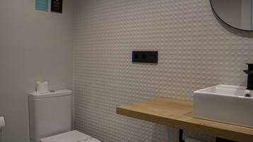 A bathroom with a white textured wall, a wooden shelf, a white sink, and a toilet.