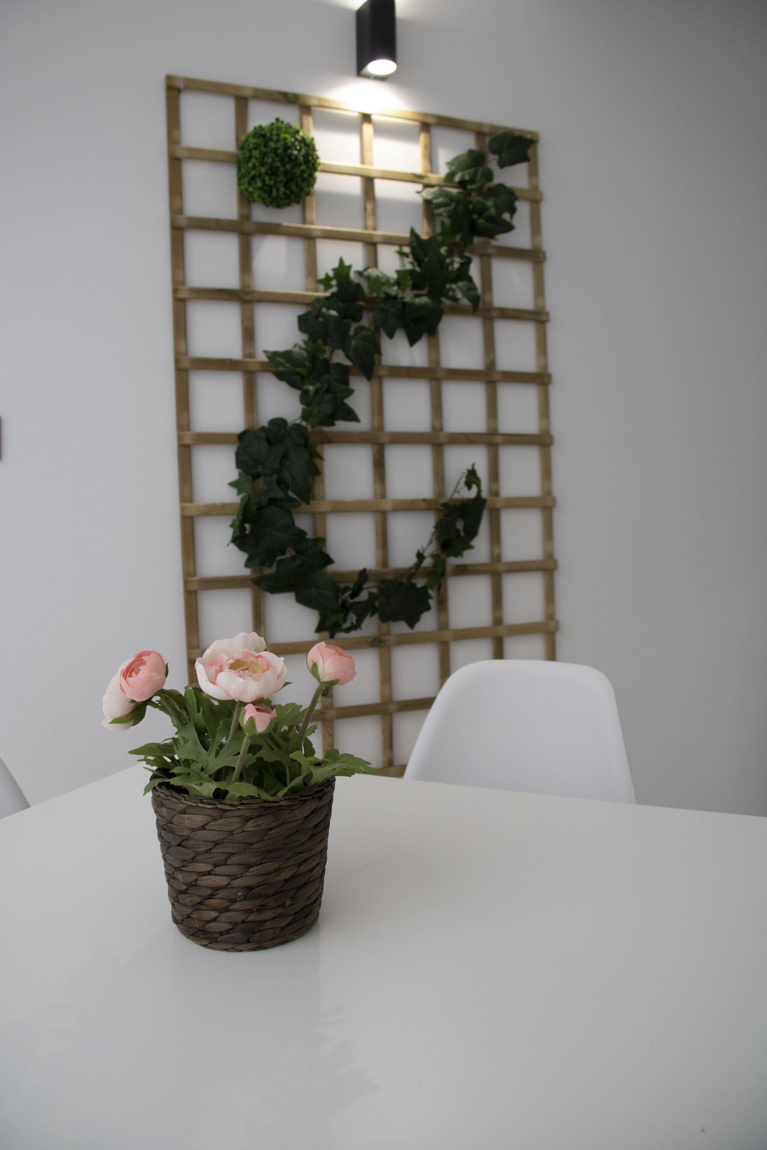 A white table with a woven basket containing pink flowers, a white chair, and a wooden lattice wall with green foliage.