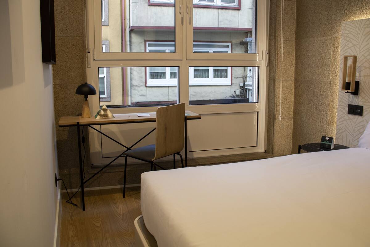 Habitación de hotel con cama, escritorio y silla, ventana con vistas a edificios y una pequeña mesa con lámpara.