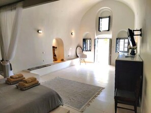 Un dormitorio amplio con una cama grande, un escritorio y una silla. Hay dos ventanas y una puerta que conduce a otra habitación.