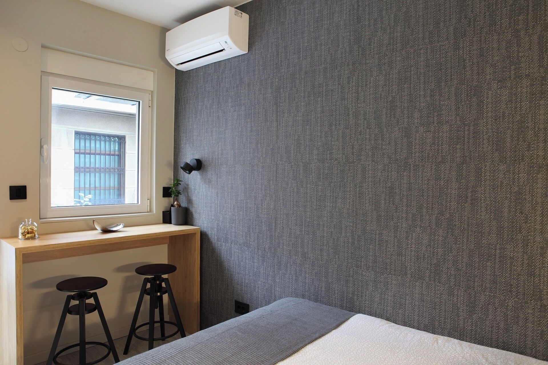A bedroom with a bed, a desk with two stools, a window with bars, and an air conditioning unit.