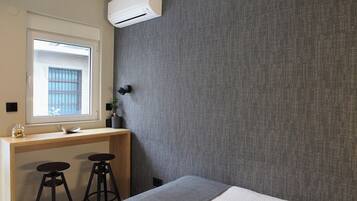 A bedroom with a bed, a desk with two stools, a window with bars, and an air conditioning unit.