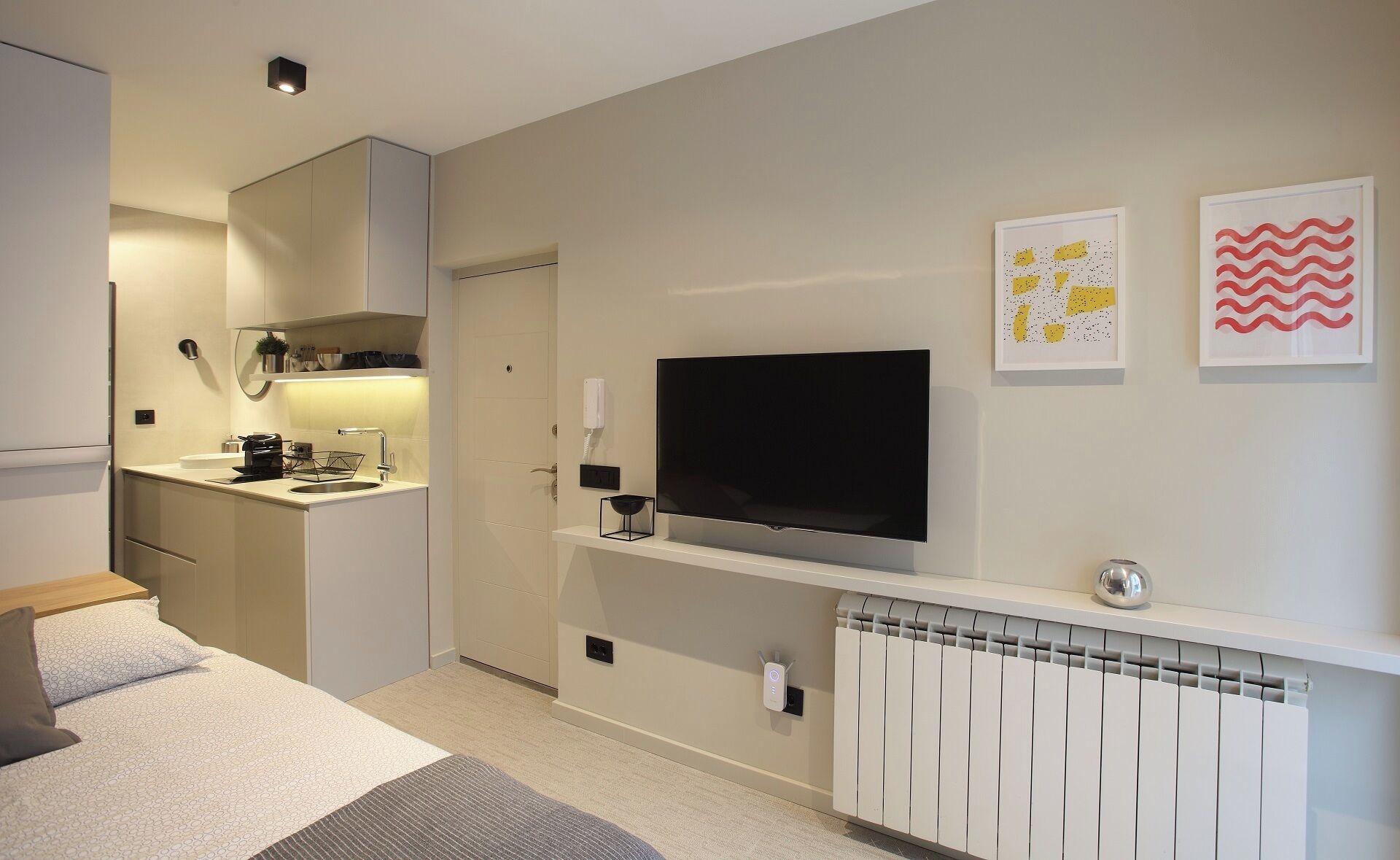 Un espace de vie moderne et compact, doté d’une kitchenette, d’un téléviseur à écran plat fixé sur une console blanche et d’un radiateur placé sous une étagère.