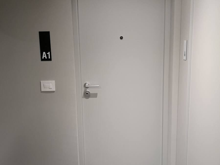 Een gesloten deur met een bordje met de tekst 'A1', een deurklink, een lichtschakelaar en een deurmat.