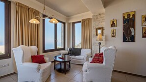 Una sala de estar con dos sillones blancos, una mesa de centro redonda y un sofá con cojines estampados. Se observan ventanas amplias con cortinas, una pared de piedra y cuadros religiosos.