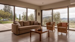 Ein geräumiges Wohnzimmer mit einem beigefarbenen Sofa, zwei Korbstühlen und einem Holztisch. Große Fenster bieten Blick auf Bäume und eine entfernte Landschaft.