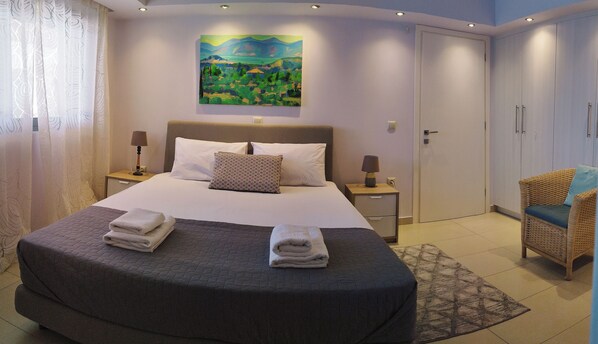 A neatly made bed with a headboard, two bedside tables with lamps, a painting on the wall, and a wicker chair.
