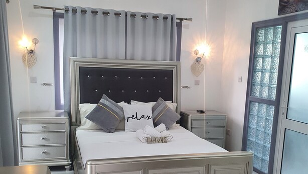 Un dormitorio moderno con una cama grande, dos mesitas de noche, un ventilador de techo y vistas al exterior a través de una ventana.