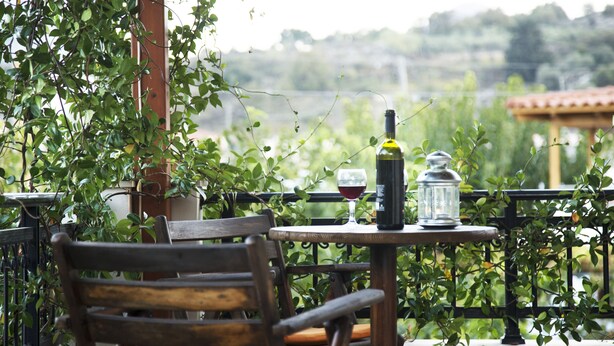 A wooden chair with a cushion, a table with a bottle and glass, and a view of greenery and buildings in the background. - Villa in a great place of Rethymno, minutes away of the most famous beaches! (Κοξαρέ)