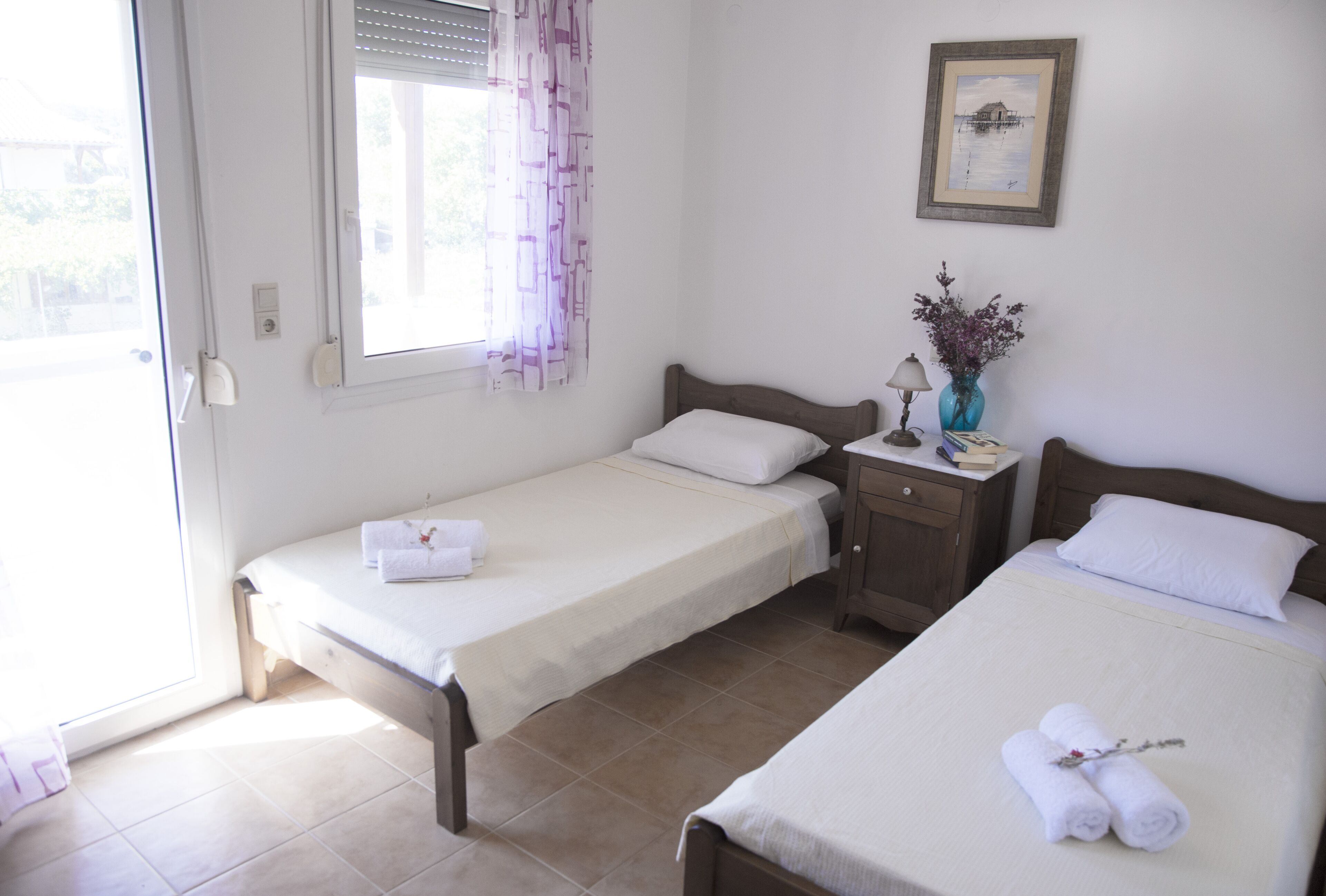 Een hotelkamer met twee eenpersoonsbedden, een klein nachtkastje met een lamp, een vaas met bloemen en een lijst met een afbeelding aan de muur.