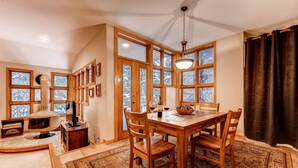 A dining area with a wooden table, chairs, and a rug. There are large windows with wooden frames, a ceiling light, and a fireplace.