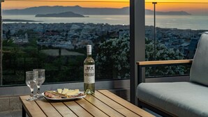 A table with a bottle of wine, glasses, and a plate of cheese and ham, overlooking a cityscape and ocean at sunset. - Karmilos Hill Villa - close to the center of Chania with private swimming pool. (Χανιά)