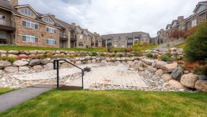 A residential complex with stone and brick buildings, a paved patio with outdoor furniture, and a landscaped garden with rocks and plants. - Bay Club Town Homes by CRMR (Frisco)
