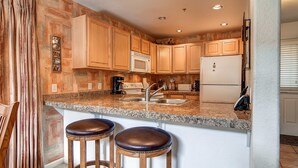 A kitchen with wooden cabinets, a granite countertop, a microwave, a refrigerator, and two bar stools. - Bay Club Town Homes by CRMR (Frisco)