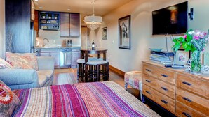 A modern bedroom with a large bed, a dresser, a TV, and a kitchen area in the background. - Cedars Lodge Condos by CRMR (Frisco)